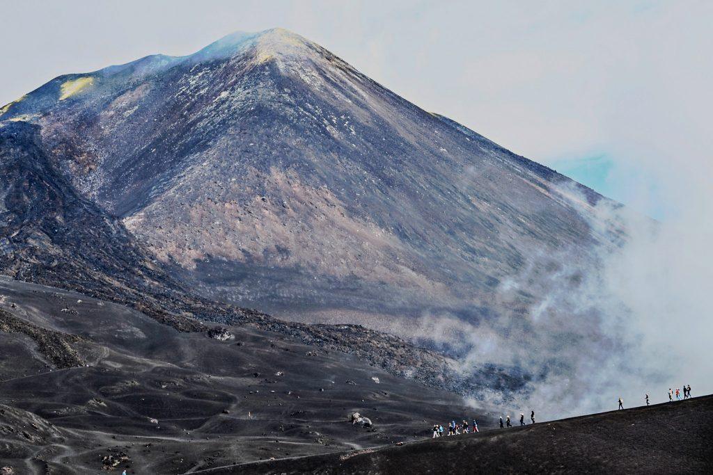 Group of hikers walking on the slopes of Mount Etna among volcanic rocks and fumaroles.