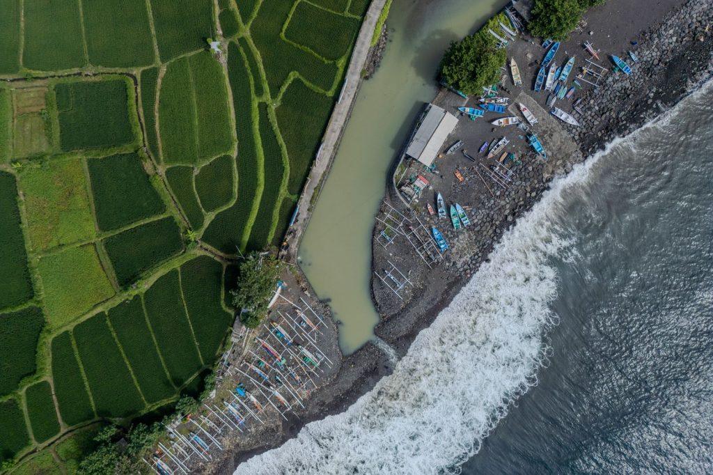 Aerial view of a Balinese coastline with fishing boats lined up near rice fields and a beach.