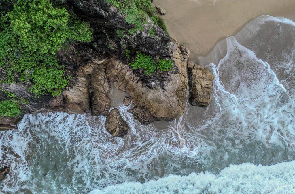 Waves crashing onto rocks and sand at the foot of a cliff covered in vegetation.