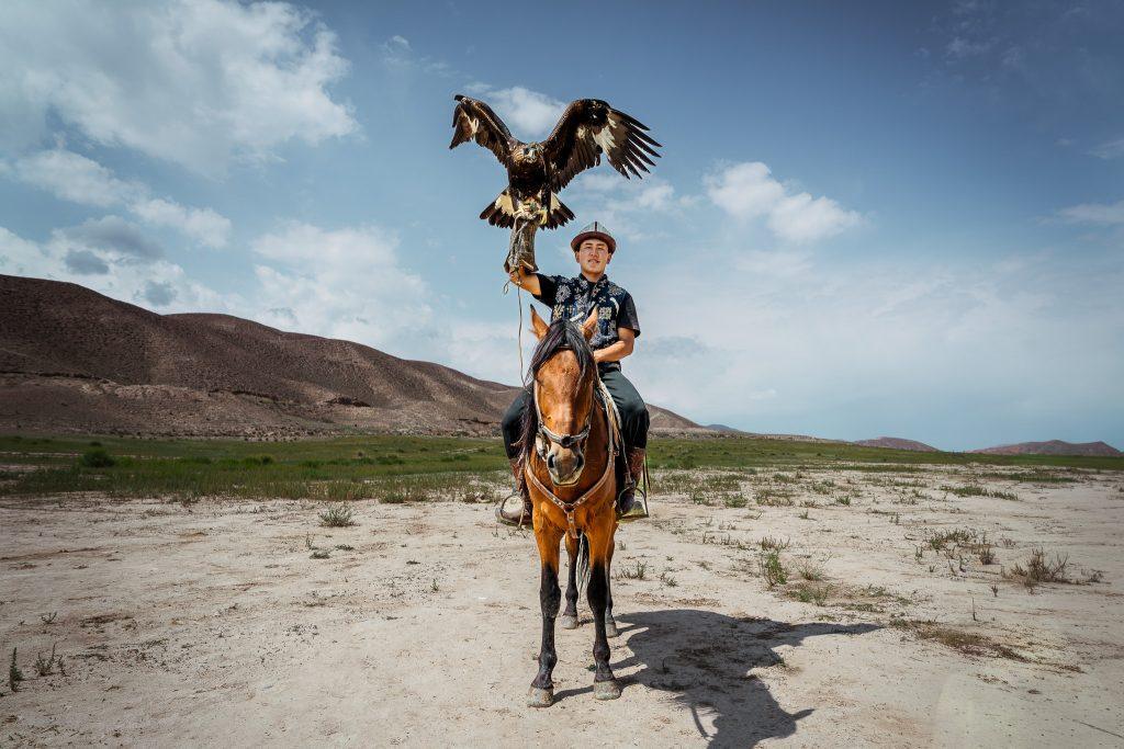 Un cavalier kirghize lève son aigle royal vers le ciel dans une plaine entourée de collines arides.