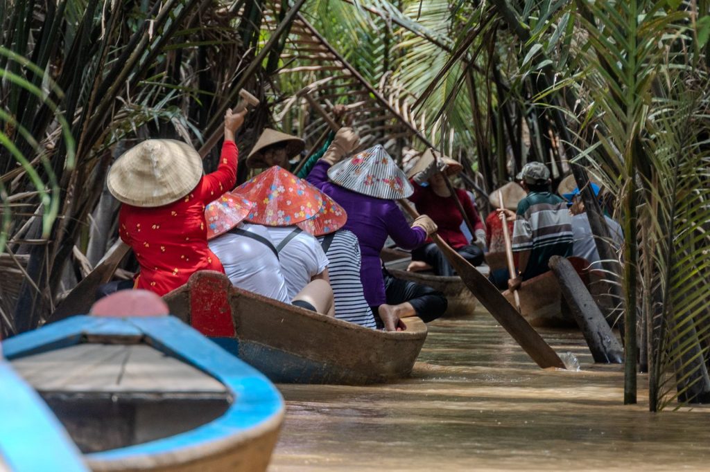 People move by boat through a narrow canal lined with palms, using paddles to navigate muddy water.