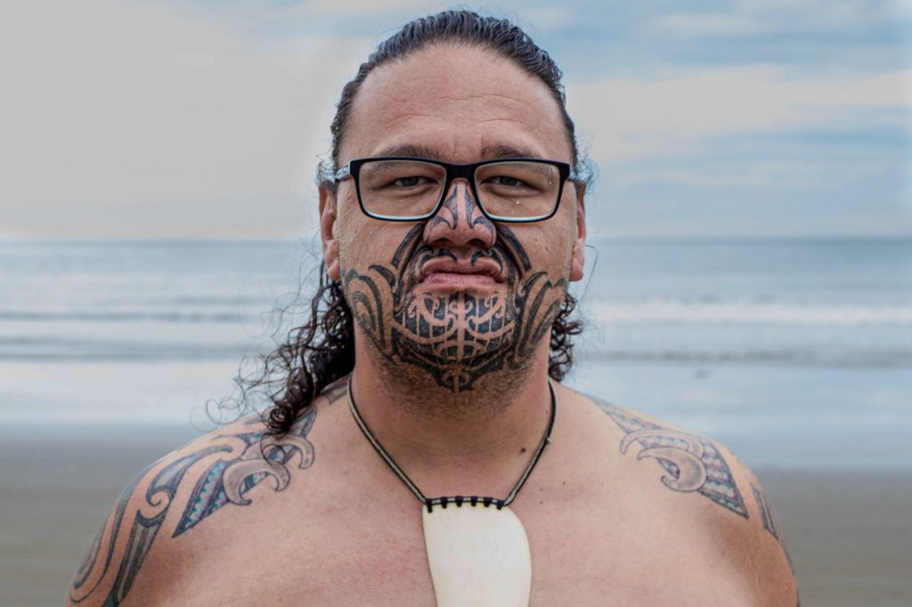 Maori man with a contemporary Ta Moko tattoo on his face standing facing the sea