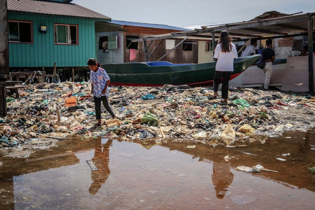 Des enfants traversent une zone couverte de déchets plastiques entre des habitations sur pilotis, au bord d'une flaque d'eau.