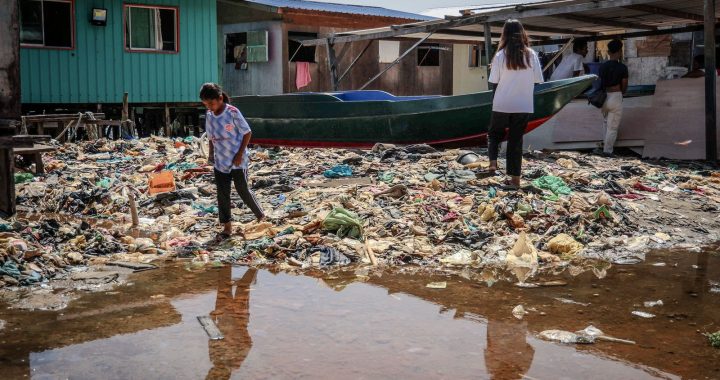 Des enfants traversent une zone couverte de déchets plastiques entre des habitations sur pilotis, au bord d'une flaque d'eau.