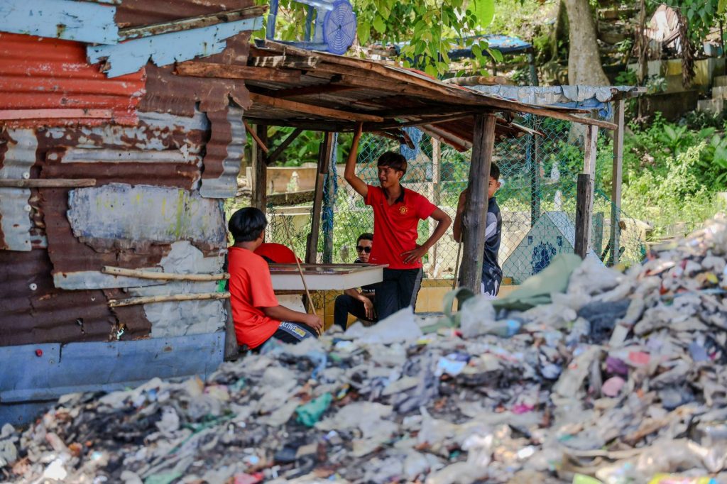 Des adolescents se retrouvent sous un abri improvisé tandis qu'un important amas de déchets occupe le premier plan dans un quartier résidentiel.