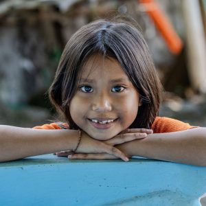 Young Balinese girl resting her head on folded arms and smiling softly towards the camera in a village setting.