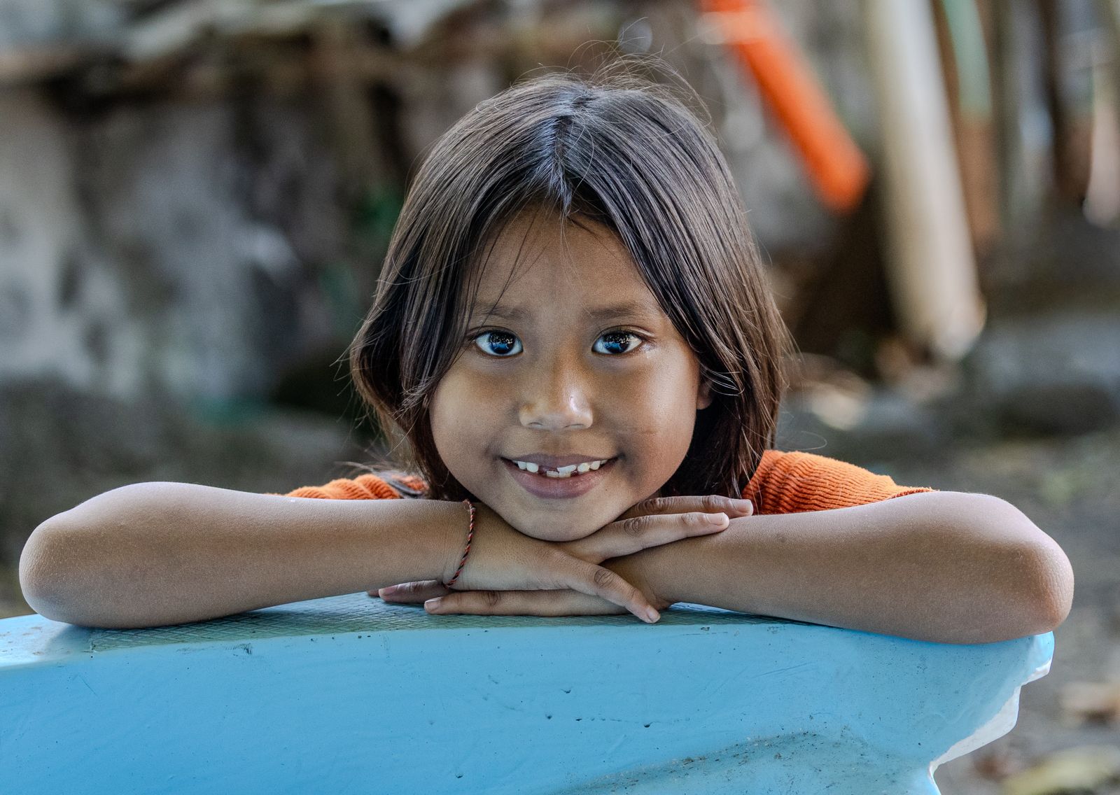 Young Balinese girl resting her head on folded arms and smiling softly towards the camera in a village setting.