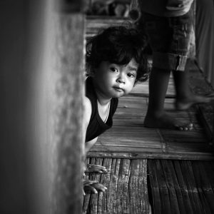 Young child peeking from behind a wooden wall in a traditional house in Java, Indonesia, black and white portrait.