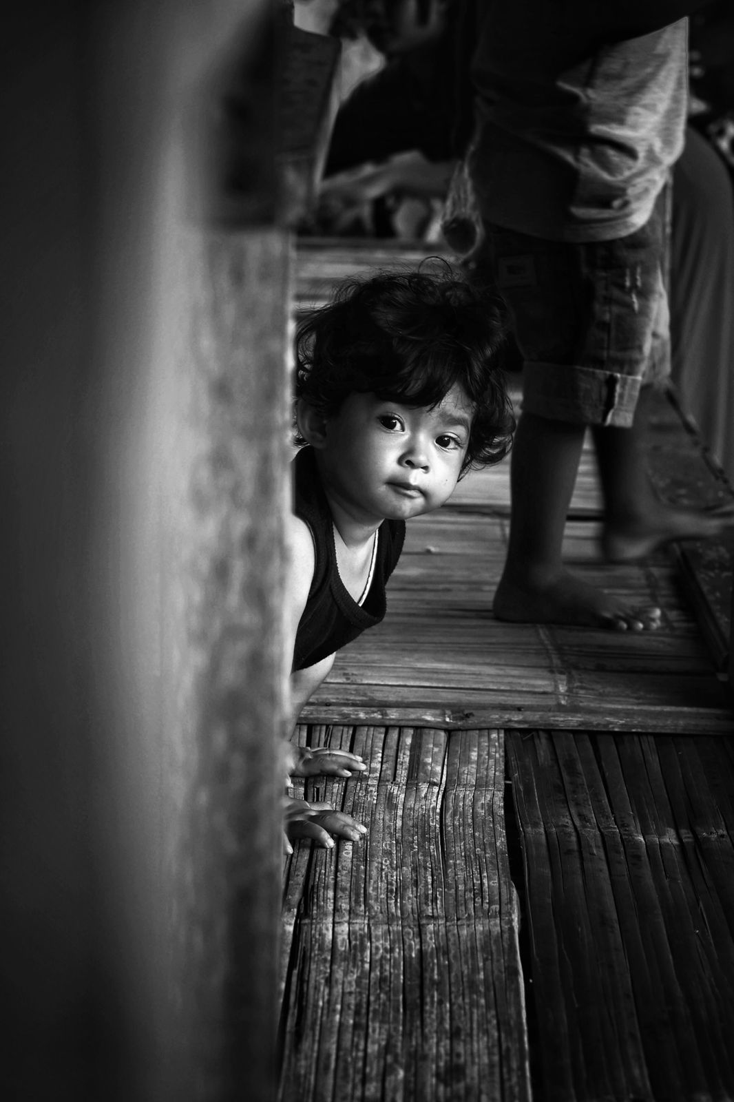 Young child peeking from behind a wooden wall in a traditional house in Java, Indonesia, black and white portrait.