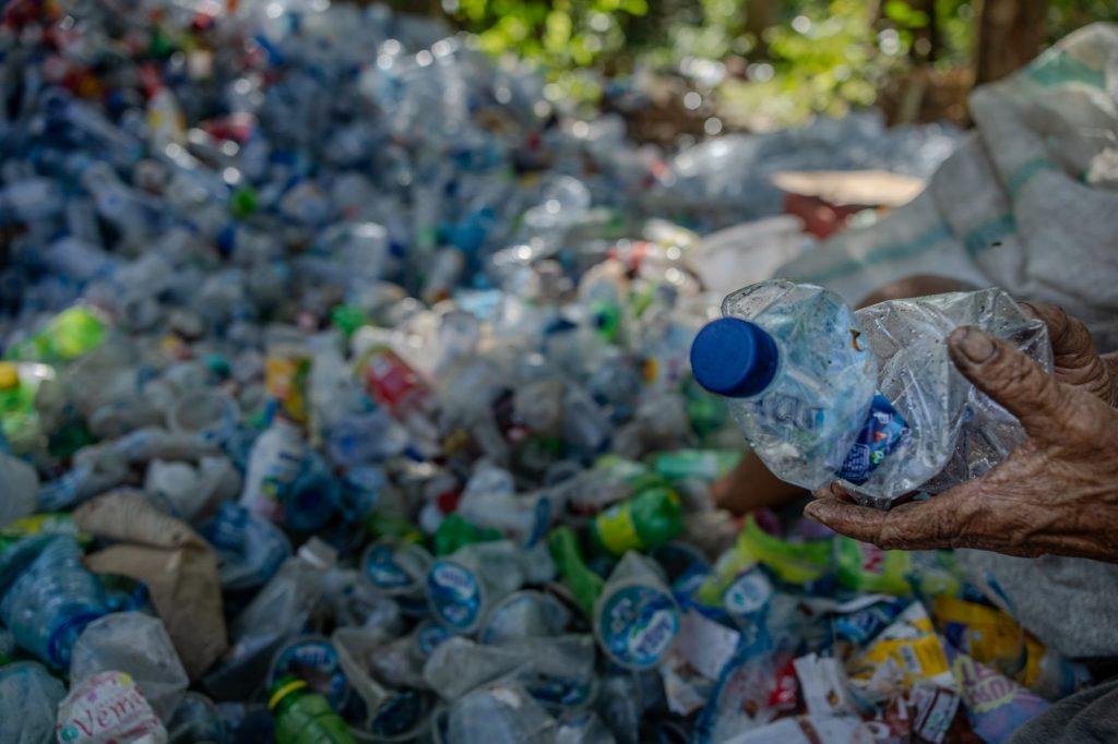 Une main tient une bouteille en plastique écrasée devant un immense amas de bouteilles et de déchets plastiques accumulés dans une décharge à ciel ouvert.