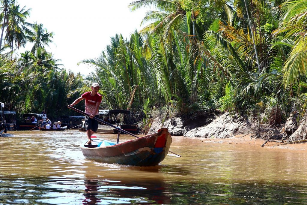 Traditional boat navigating a tropical river surrounded by palm trees in Southeast Asia.