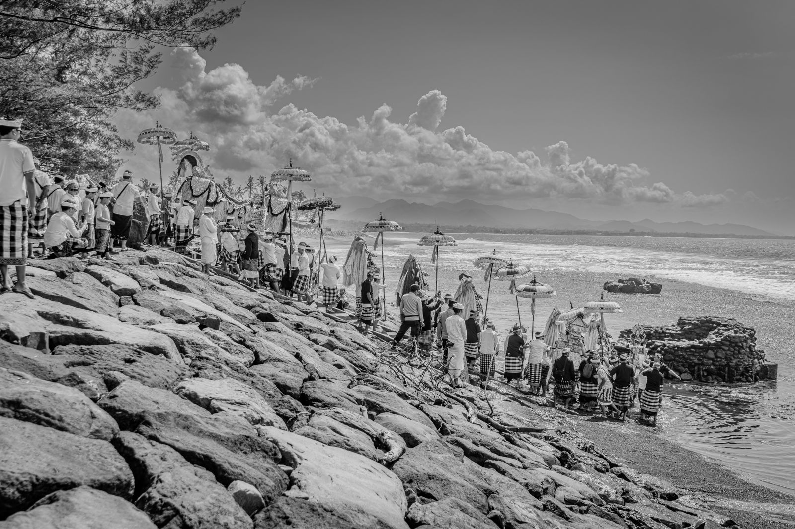 Balinese ceremonial procession descending to the sea beneath traditional umbrellas along the coast of Bali.