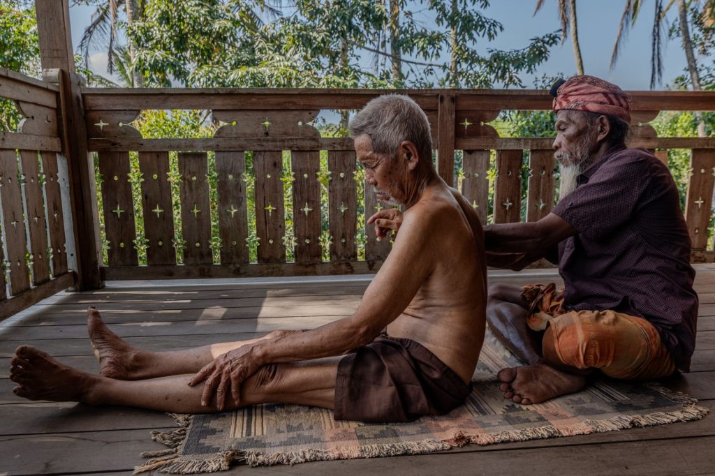 A balian treats a man sitting on a mat on the wooden terrace of a Balinese house.