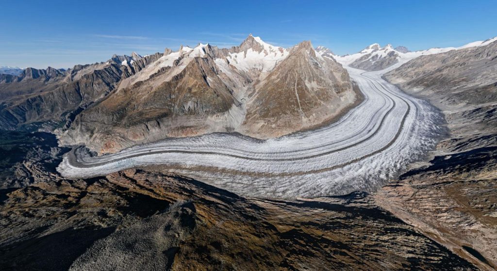 Aerial view of the Great Aletsch Glacier in the Swiss Alps, showing its curved ice flow, dark moraines and surrounding rocky mountains. Credit : © Rémih / Wikimedia Commons